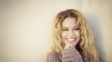 Hymyilevä sisustussuunnittelija Milla Alftan pitkissä vaaleissa hiuksissaan beigen seinän edustalla