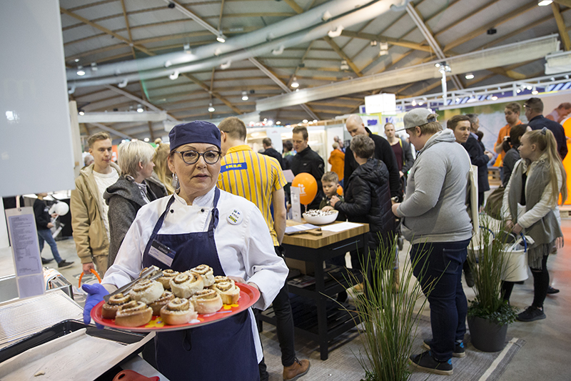 Ikean työntekijä käsissään lautasellinen korvapuusteja, taustalla messuyleisöä