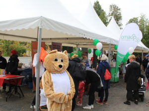 Nalle pukuinen henkilö teltan edessä tapahtumavierailijoiden kävellessä taustalla