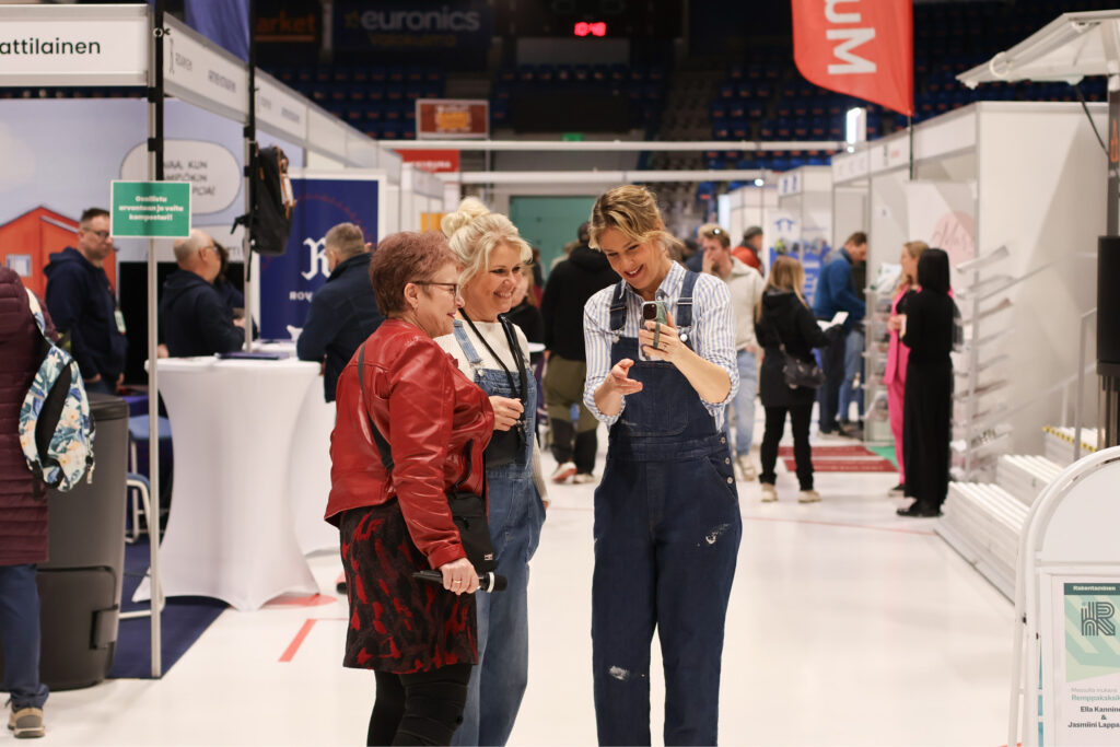 Ella Kanninen näyttää iloisena valokuvaa puhelimeltaan punaisessa nahkatakissa olevalle rouvalle Rakentajamessuilla